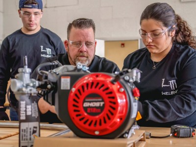 Three people, a teacher and two students, work on a wooden table next to a red circular saw. Each person wears clear safety glasses.
