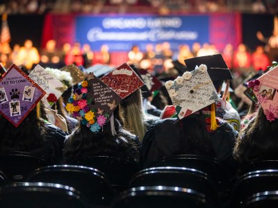 A group of graduates is seated with their decorated caps visible, featuring colorful designs, personal messages, and cultural symbols. In the background, a stage and a large banner reading "Chicano Latino Commencement Celebration" are illuminated, surrounded by people dressed in academic regalia.