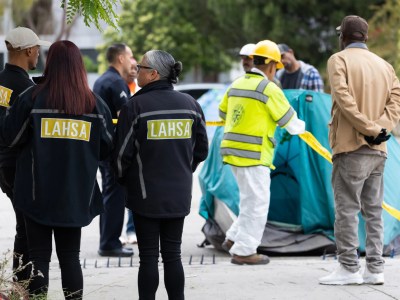 LAHSA workers and others in high-visibility gear stand near a tent, surrounded by yellow caution tape. The scene appears to involve a city operation, possibly addressing a homeless encampment. People in the background are observing or engaged in conversation.