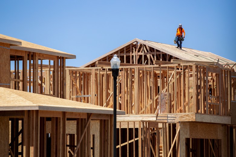 A wide view of houses under construction with wooden framing. A construction worker wearing a bright orange shirt can be seen standing in the roof of one of the houses.