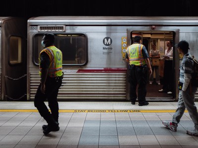 Two workers in yellow and orange safety vests stand on an underground subway platform. One worker speaks to two people on a train, as the doors remain open. Another person walks by and observes the conversation. The train remains stopped at the station.