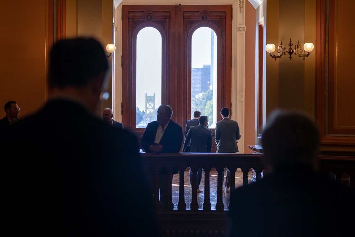 A group of individuals is gathered near large, arched windows in a dimly lit hallway. The silhouettes of a few people stand against the bright backdrop of the windows, which frame a view of a cityscape and a bridge in the distance. The setting appears to be inside a government building, with elegant woodwork and soft lighting from wall-mounted lamps. The scene evokes a sense of quiet anticipation or reflection, with people standing in conversation or gazing out the windows.