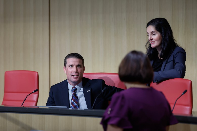 State Senate President Pro Tempore Mike McGuire, wearing a tie and blue suit is seated in a red chair at an elevated dias in a hearing chamber. Next to and in front of him, two people stand in conversation.