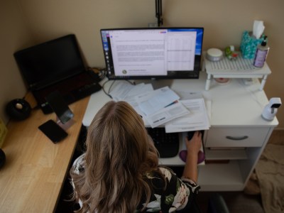 An overhead view of a person with blonde hair sitting in a chair looking at a computer on a desk filled with various papers.