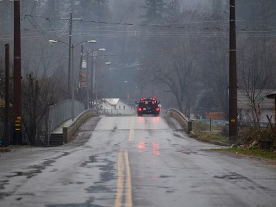 A truck drives over a bridge and down a wet road. The rear of the truck is visible and the brake lights are engaged. The setting is a small, rural, town. The weather is drizzly, foggy and grey.