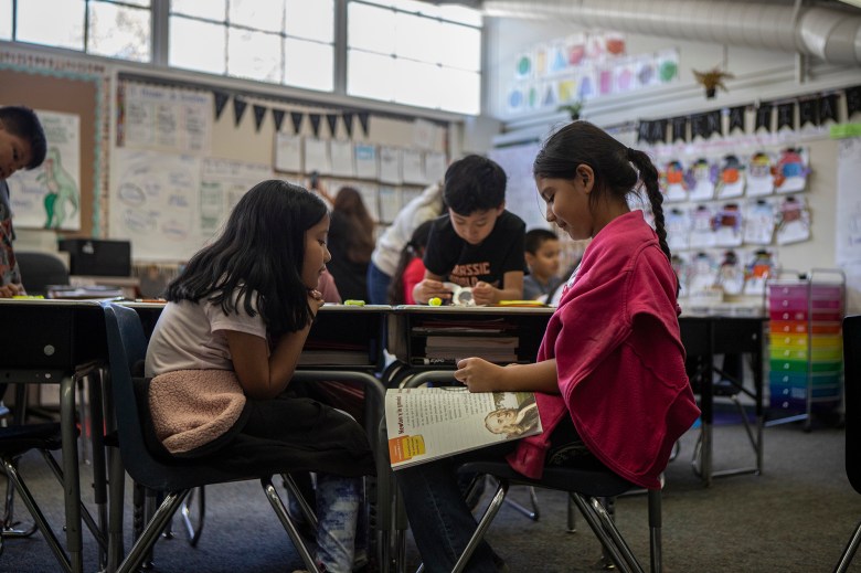 A close-up view of two young students as they sit in chairs facing each other. The student on the right wears a hot pink sweater while looking down at a workbook that rests on her thighs. The student on the left wears a light pink shirt and rest her hands on her chin.