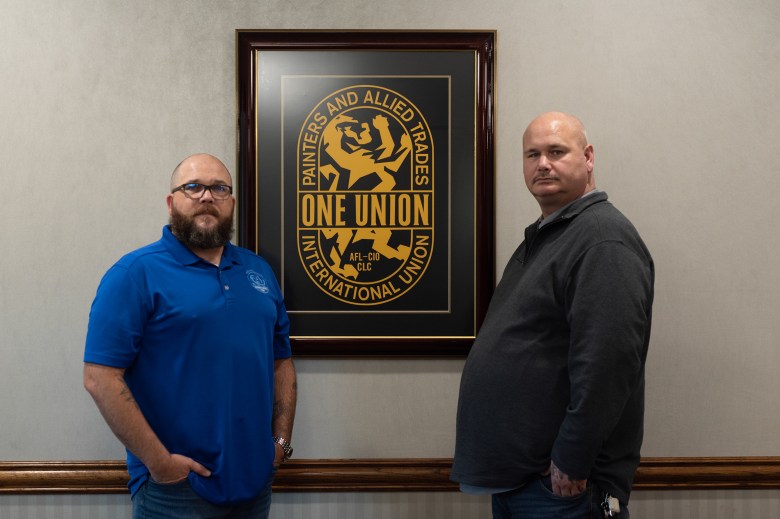 Two people stand in front of a framed International Union of Painters and Allied Trades logo hung up on a wall in an office. The person on the left