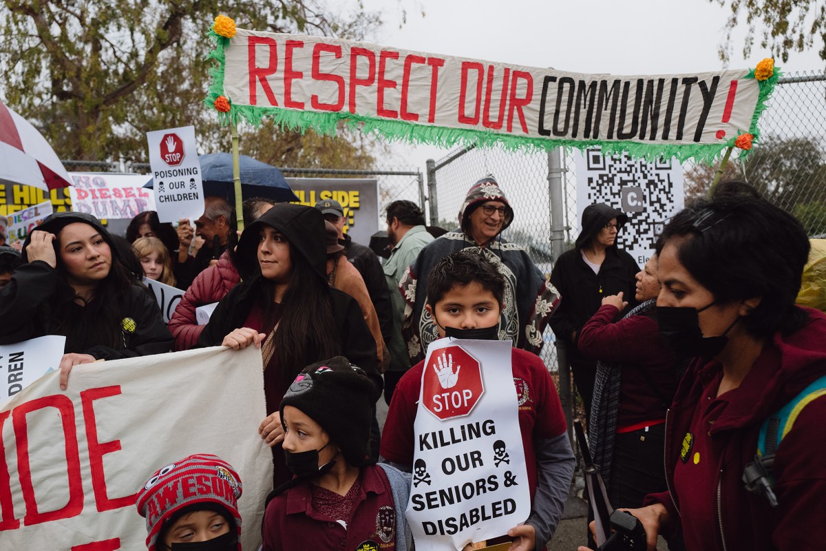 People, including young children, gather during a rally and hold signs that read "stop killing our senior disabled" and "stop poisoning our children." Two people in the back hold a banner that reads "respect our community!"