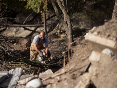 A person in working overalls uses a chainsaw to cut into a tree trunk in a park. Debris from concrete slabs and a chopped-down tree surround the person as they work in a park.