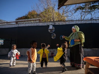 A person, wearing a green hijab and blouse with a long black skirt, uses a bucket and a ring device to release bubbles to a group of children playing nearby.