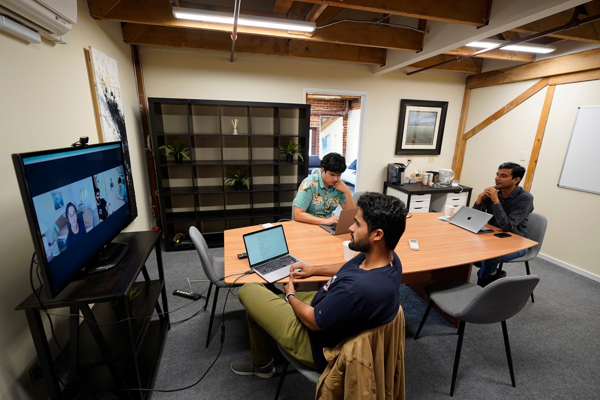 Three people sit around a table in an office meeting room with laptops in front of them. One person is looking at their computer while the other two look at a television screen with two people other people in a video meeting call.