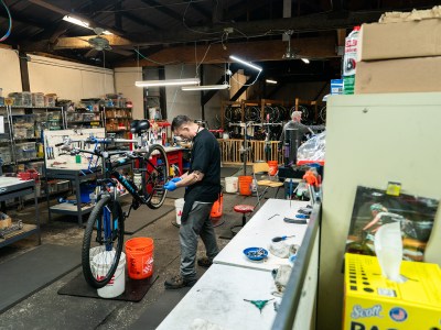 A person wearing blue gloves working on the back tire of a bicycle that is being held up in the center of a workshop. The workshop has buckets on the ground and tables with different tools around the edges, and bike racks with bicycles in the background.