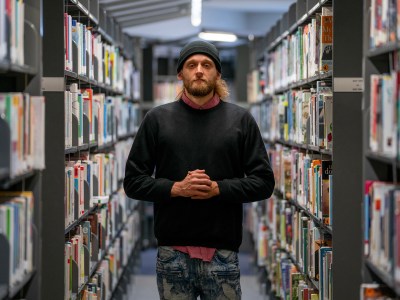 A person stands centered in the aisle of a library, surrounded by shelves full of books. They wear a black beanie, black sweater, and distressed jeans. Their hands are clasped together in front of them, and they gaze directly into the camera with a neutral expression. The bookshelves extend into the background, creating depth.