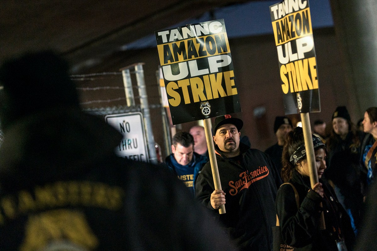 People in sweaters and warm clothing walk with picket signs on signs outside the fence of an Amazon Warehouse. The signs the people are holding say, “ TANNC Amazon ULP Strike.”