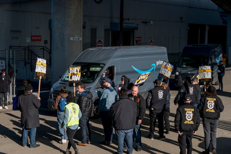 People in sweaters and warm clothing walk with picket signs on signs outside an Amazon warehouse near an Amazon van parked in front of them. The signs the people are holding say, “ TANNC Amazon ULP Strike.”