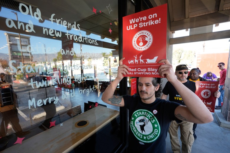 A line of people holding signs and marching in front of a Starbucks coffee shop window. The person in front of the line has a red sign over their head saying, “We’re on ULP Strike!”