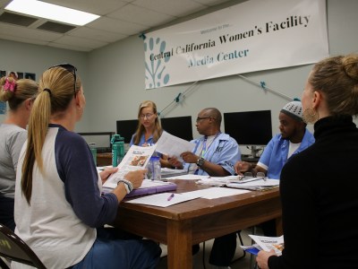 A small group of individuals sits around a table in a classroom-like environment, engaged in discussion and reviewing printed materials. A banner on the wall reads "Central California Women's Facility Media Center." Behind the participants, computer monitors are arranged on desks. The atmosphere appears collaborative and focused, with participants exchanging ideas and referencing documents.