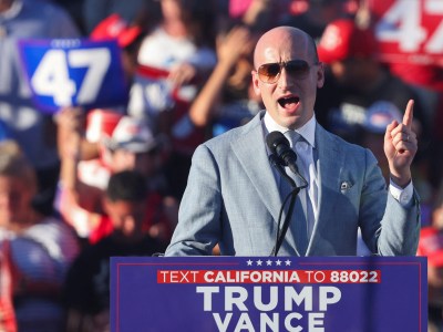 A person in a gray colored suit at a podium labeled “ TEXT CALIFORNIA TO 8802 TRUMP VANCE” talking into a microphone on a stage. Behind them is a crowd of people, with one person holding a blue sign with the number 47 on it.